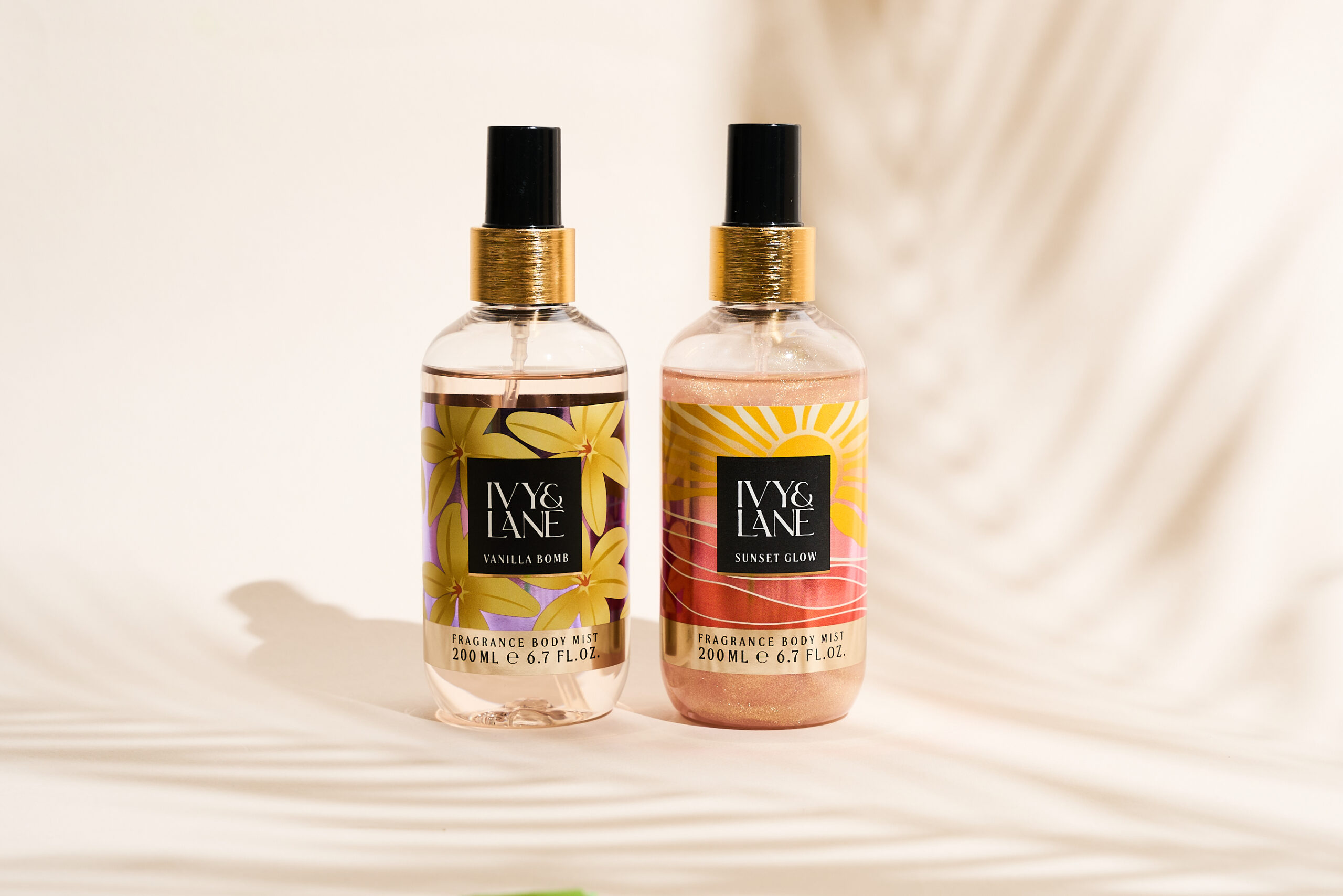 Der Bodymist IVY & LANE Vanilla Bomb und IVY & LANE Sunset Glow stehen nebeneinander vor einem weißen Hintergrund mit dem Schatten eines Palmenblatts im Hintergrund.
